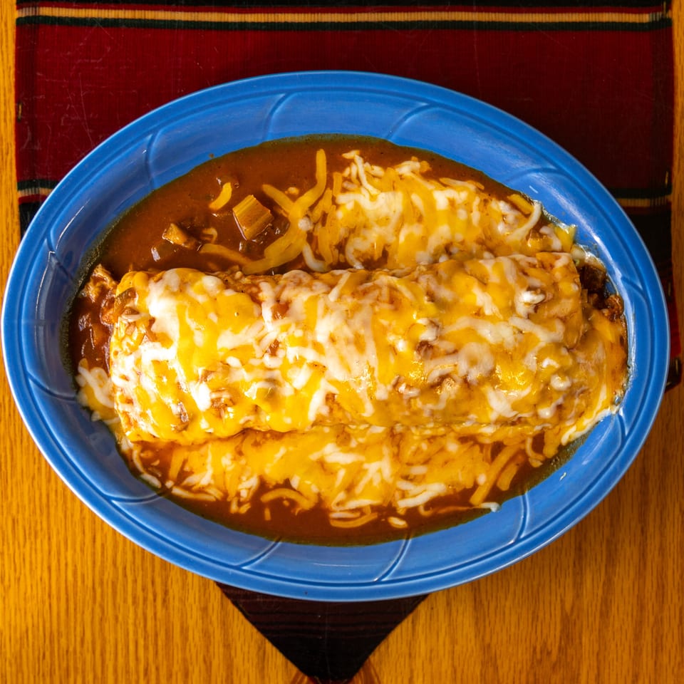 Cheese-covered enchiladas in red sauce served on a blue plate over a colorful woven placemat.