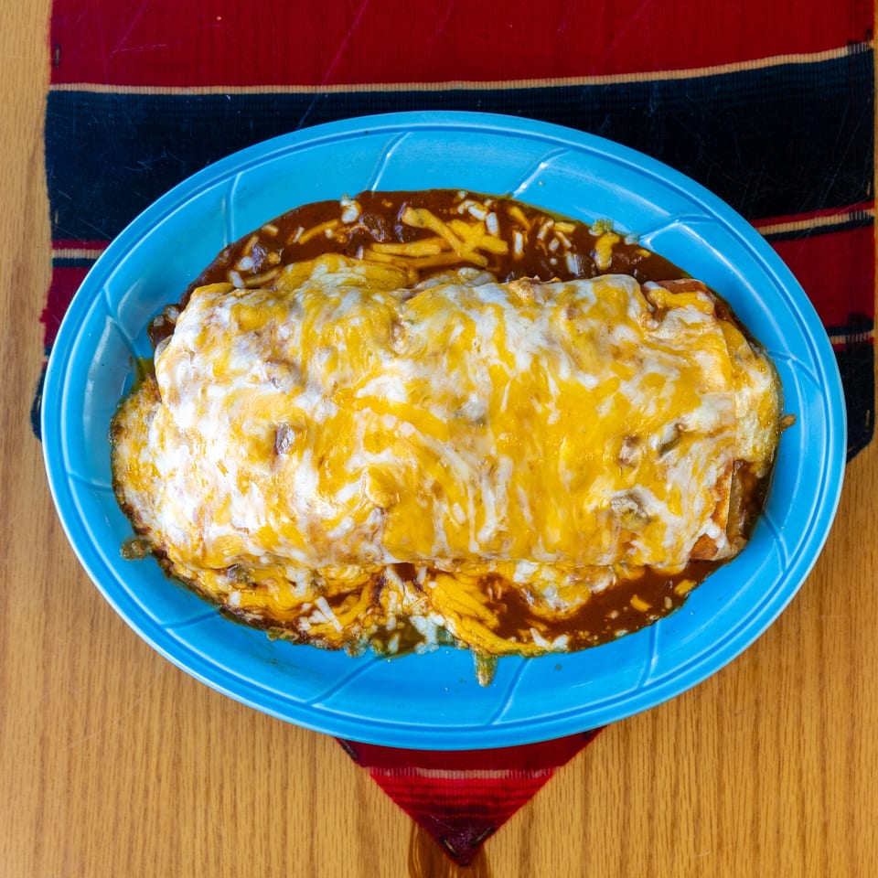 Cheese-covered enchiladas on a blue plate, served with fries, brown sauce, and chopped onions on a wooden table.