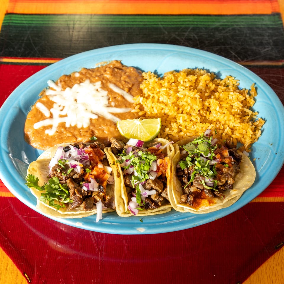Blue platter of three beef tacos with cilantro and onions, Mexican rice, refried beans, lime, and tortillas on a colorful tablecloth.