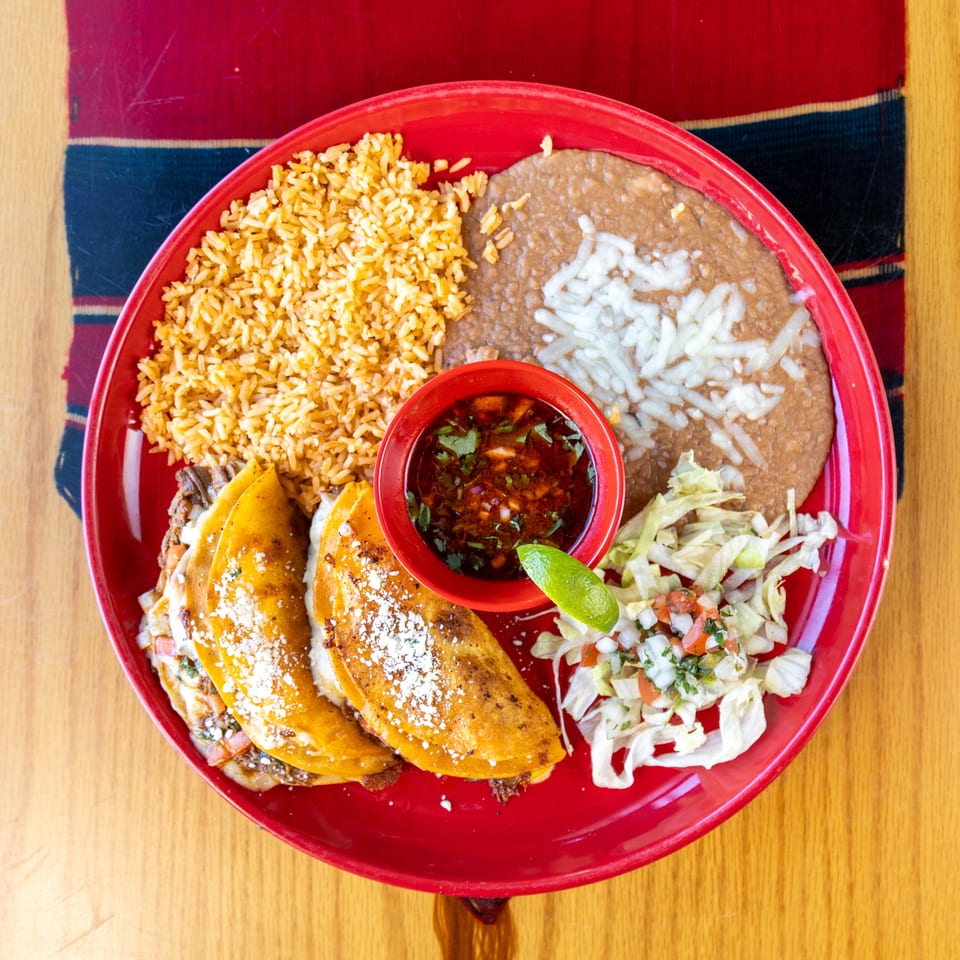 Overhead view of a Mexican platter with birria tacos, rice, refried beans, salsa, lime, and cabbage salad on a red plate.
