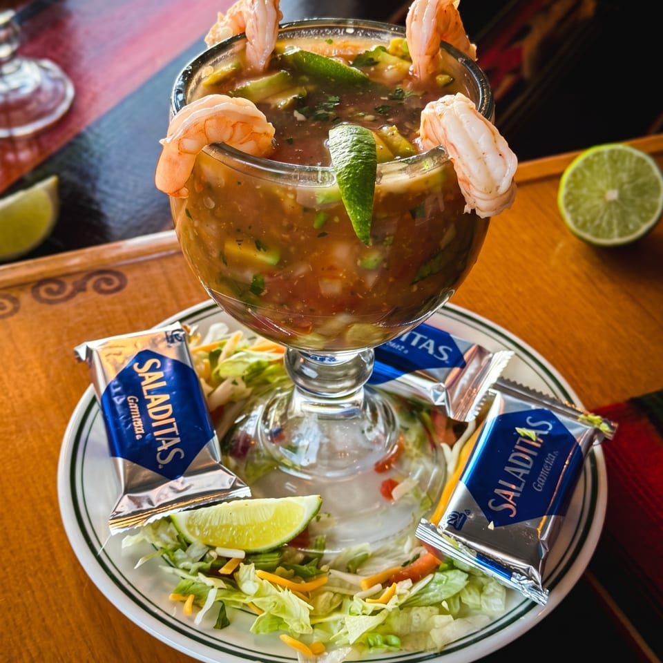 Shrimp cocktail in a glass goblet garnished with lime, surrounded by salad, lime wedges, and Saladitas crackers on a plate.