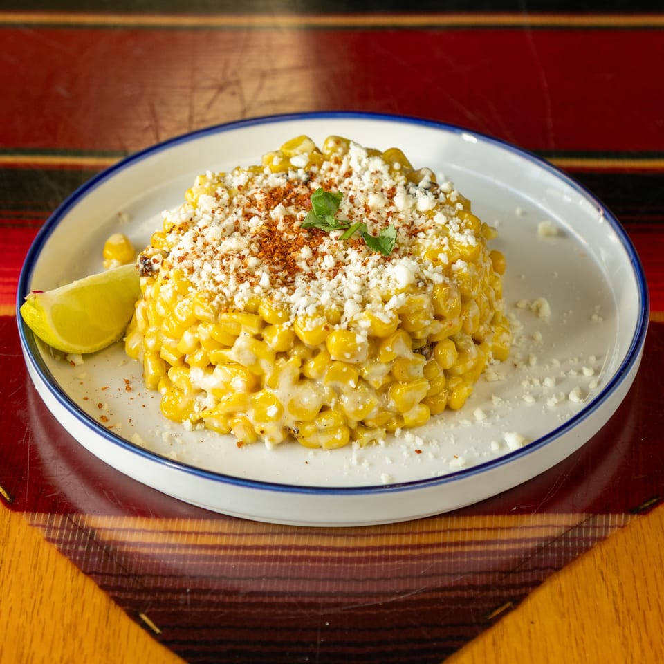 Creamy roasted corn topped with crumbled cheese, chili seasoning, and herbs, served with a lime wedge on a white plate.
