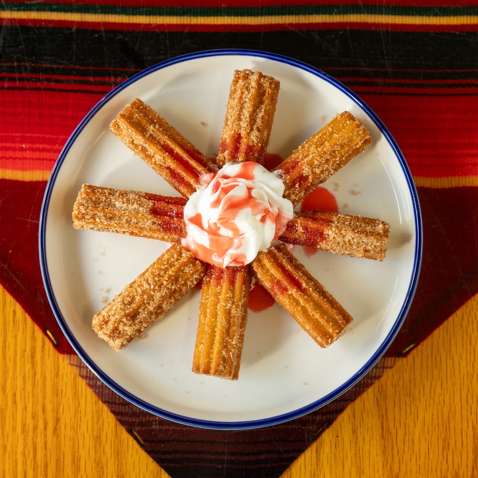 Cinnamon-sugar churros arranged around whipped cream and strawberry sauce on a plate atop a colorful woven tablecloth.